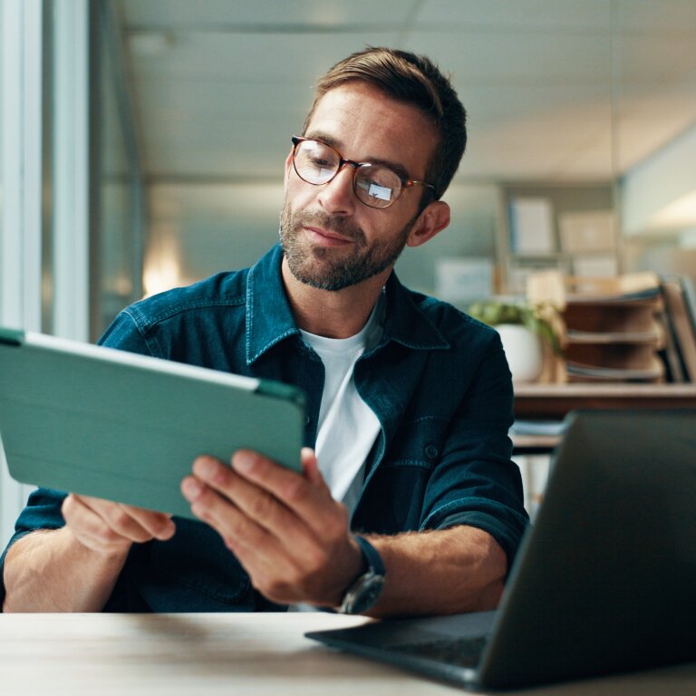 A man in a dark shirt sits at an office desk and studies documents on a tablet, with an open laptop in front of him