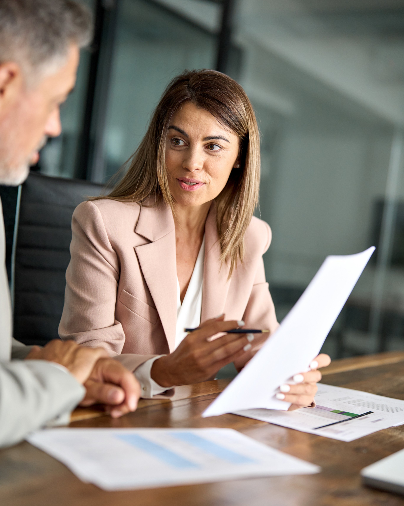 A woman in a pale blazer holding a document and discussing it with a colleague at a wooden meeting table
