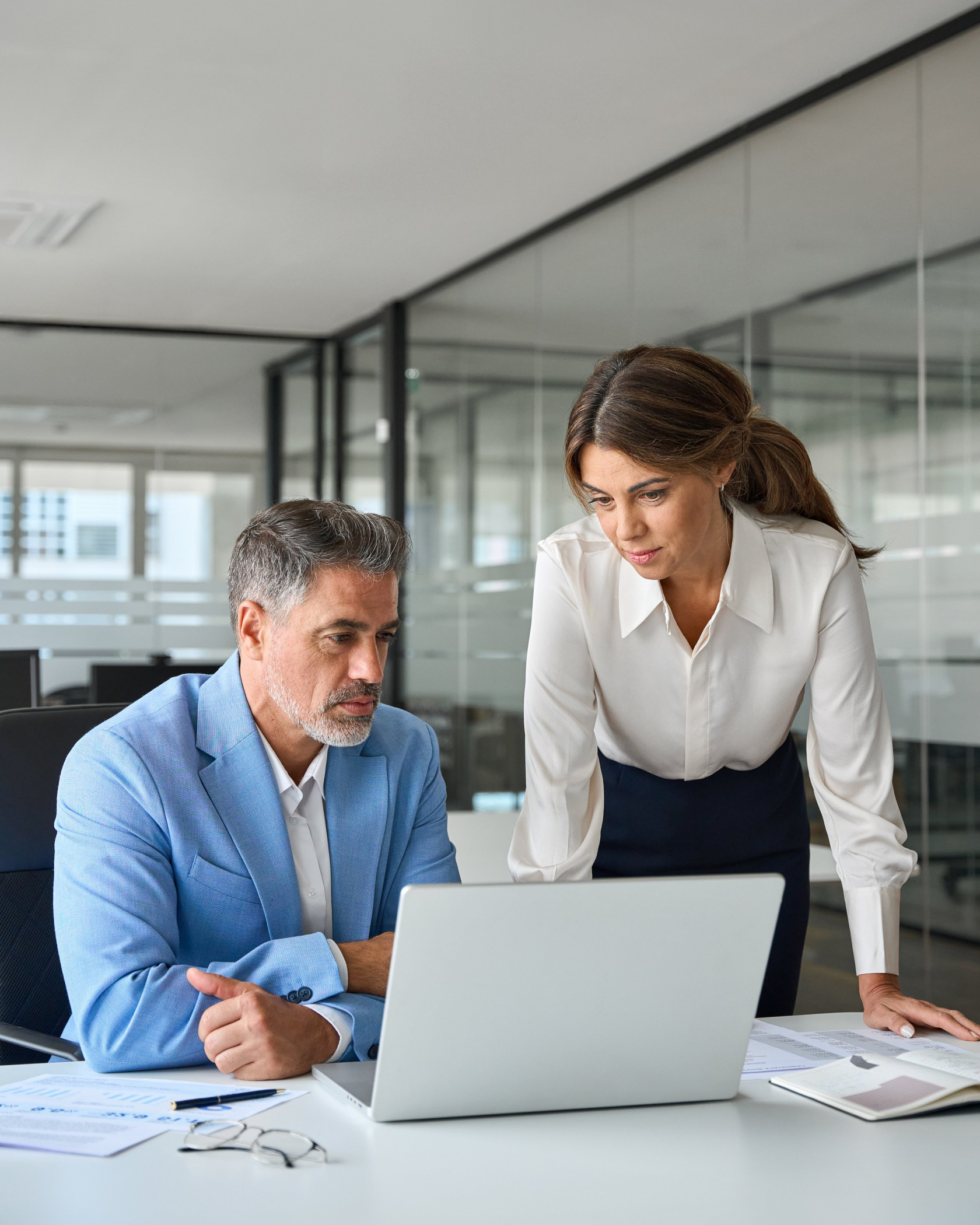 A man and a woman at a white desk looking together at a laptop screen in an open-plan office