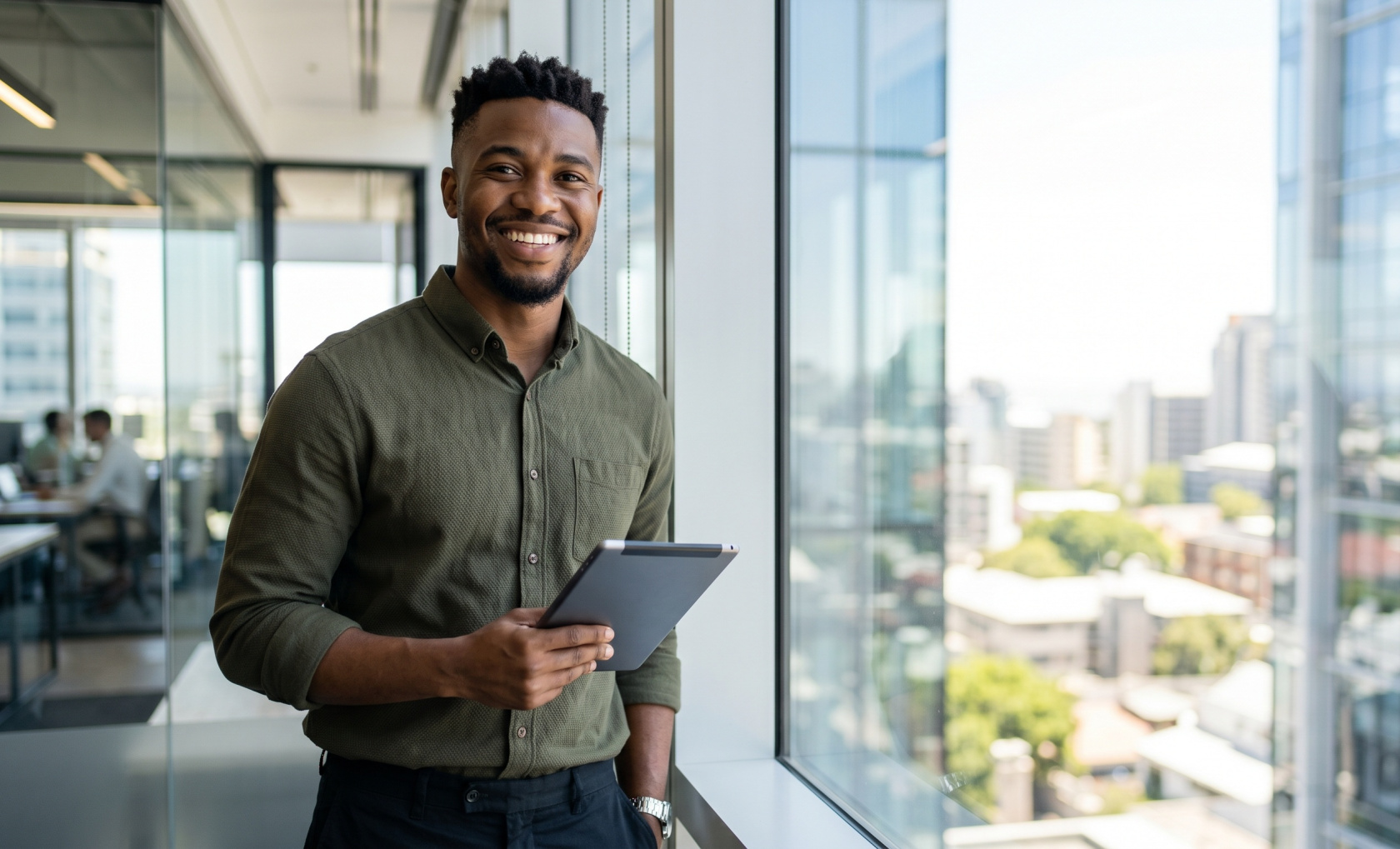 A smiling man in an olive shirt stands with a tablet at an office window, colleagues working behind a glass wall in the background