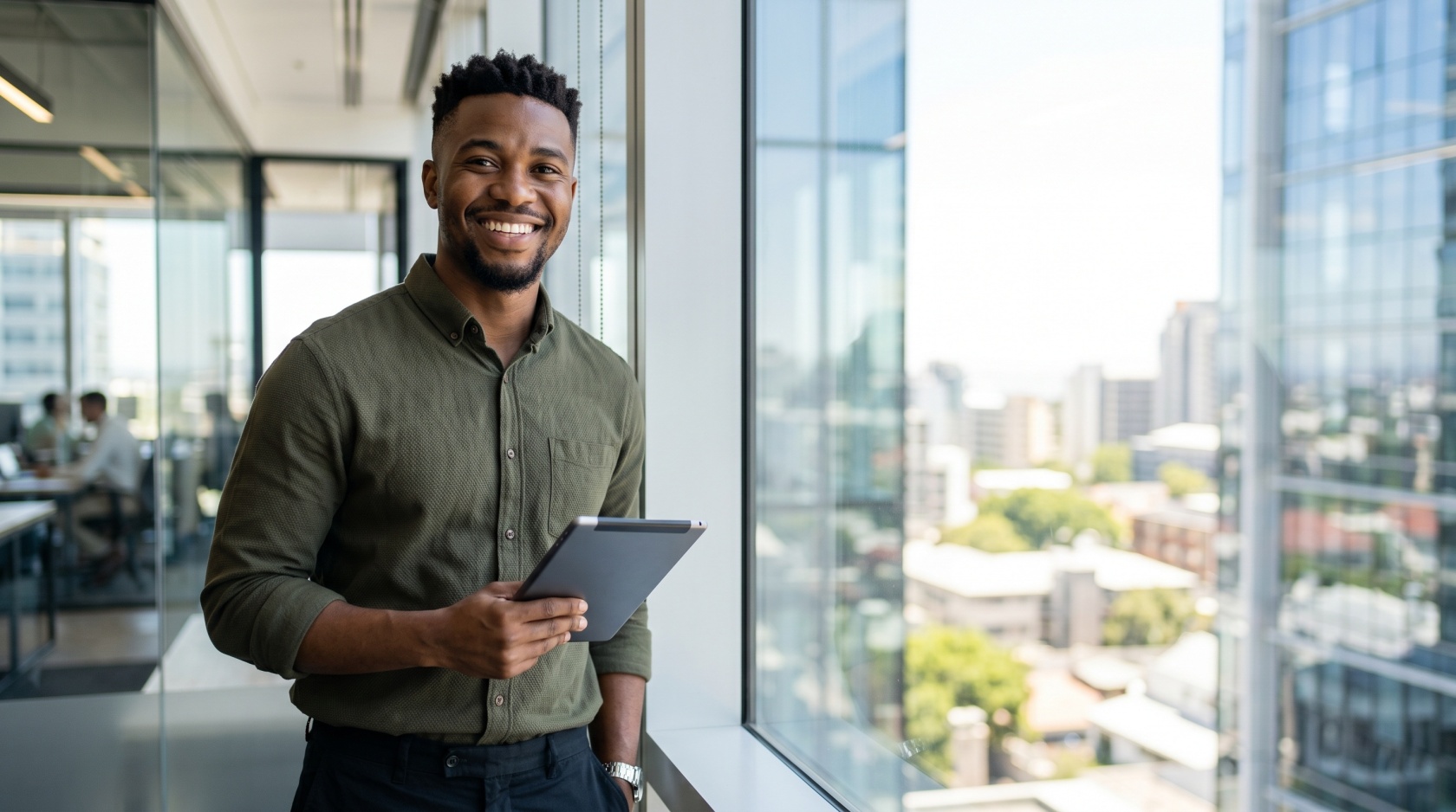 A smiling man in an olive shirt stands with a tablet at an office window, colleagues working behind a glass wall in the background