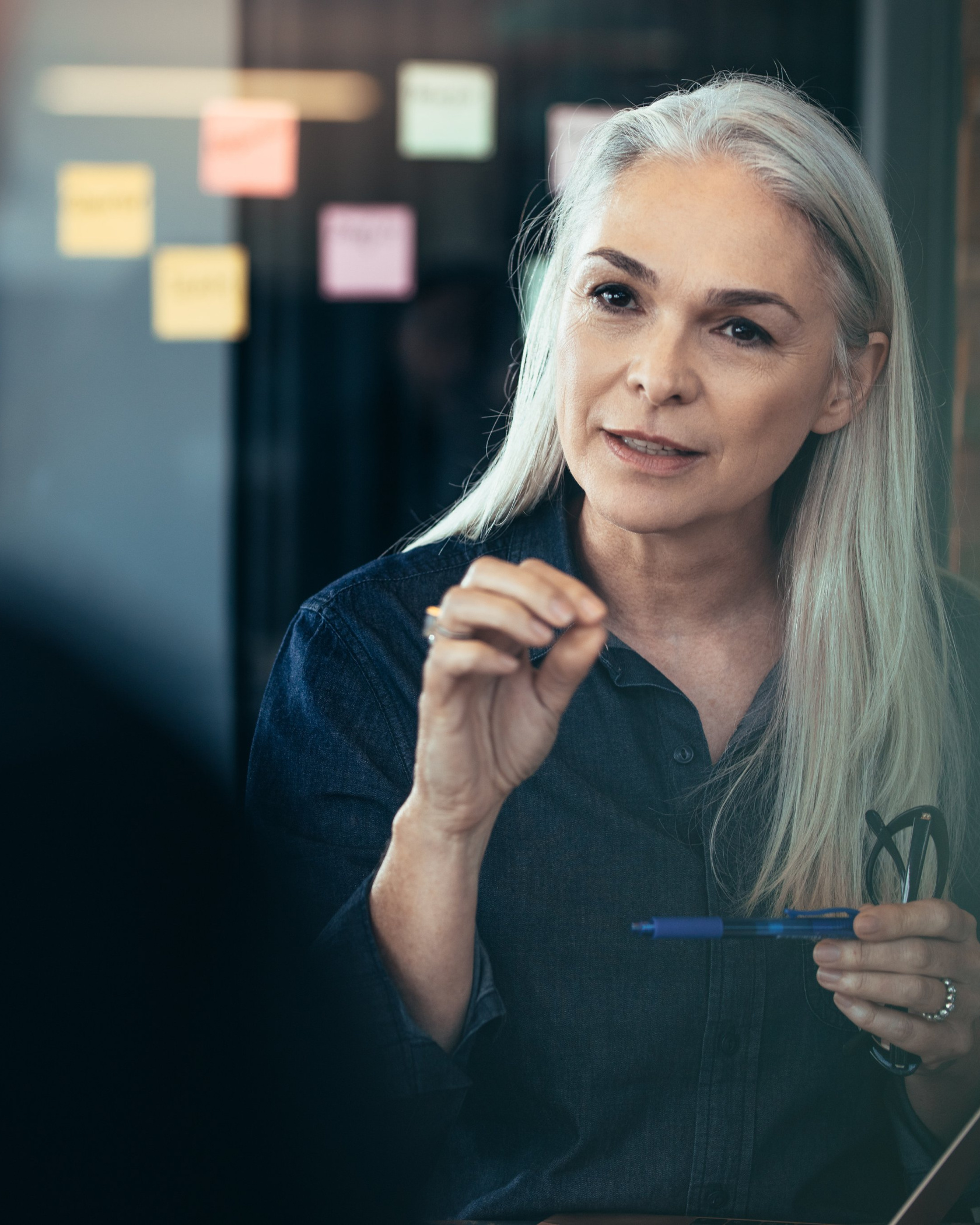 A woman with long silver hair speaking and gesturing, a pen in her other hand, in front of a glass wall covered with sticky notes