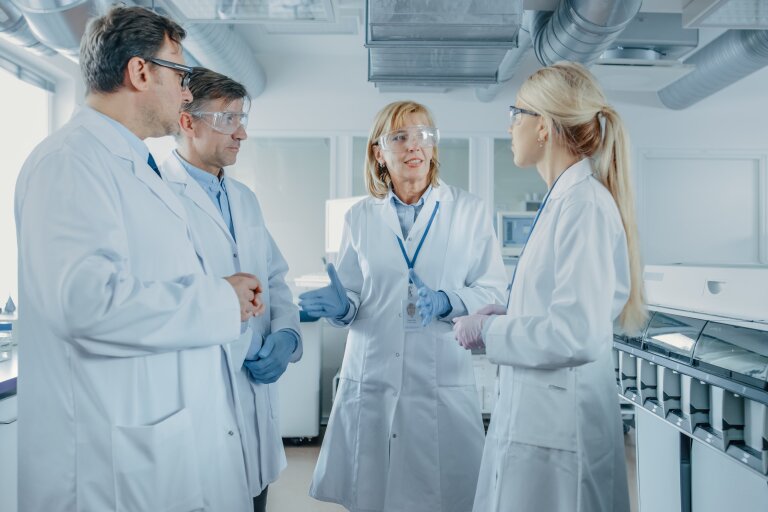 Four researchers in white coats and safety glasses talking together in a laboratory