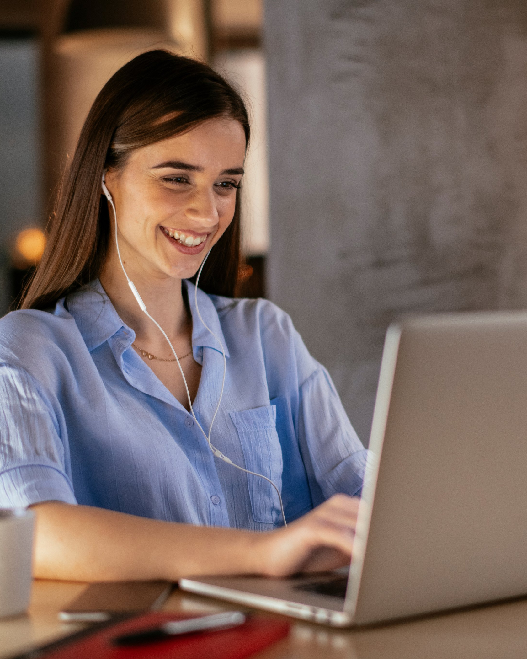 A smiling woman wearing earphones types on a laptop at a table, a phone and a notebook lying beside her