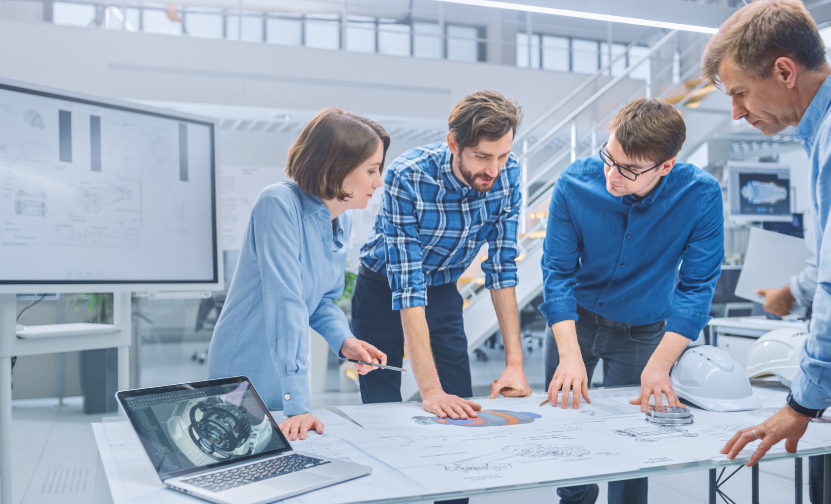 Four engineers leaning over technical drawings on a glass table in a bright industrial hall, a laptop showing a 3D model beside them