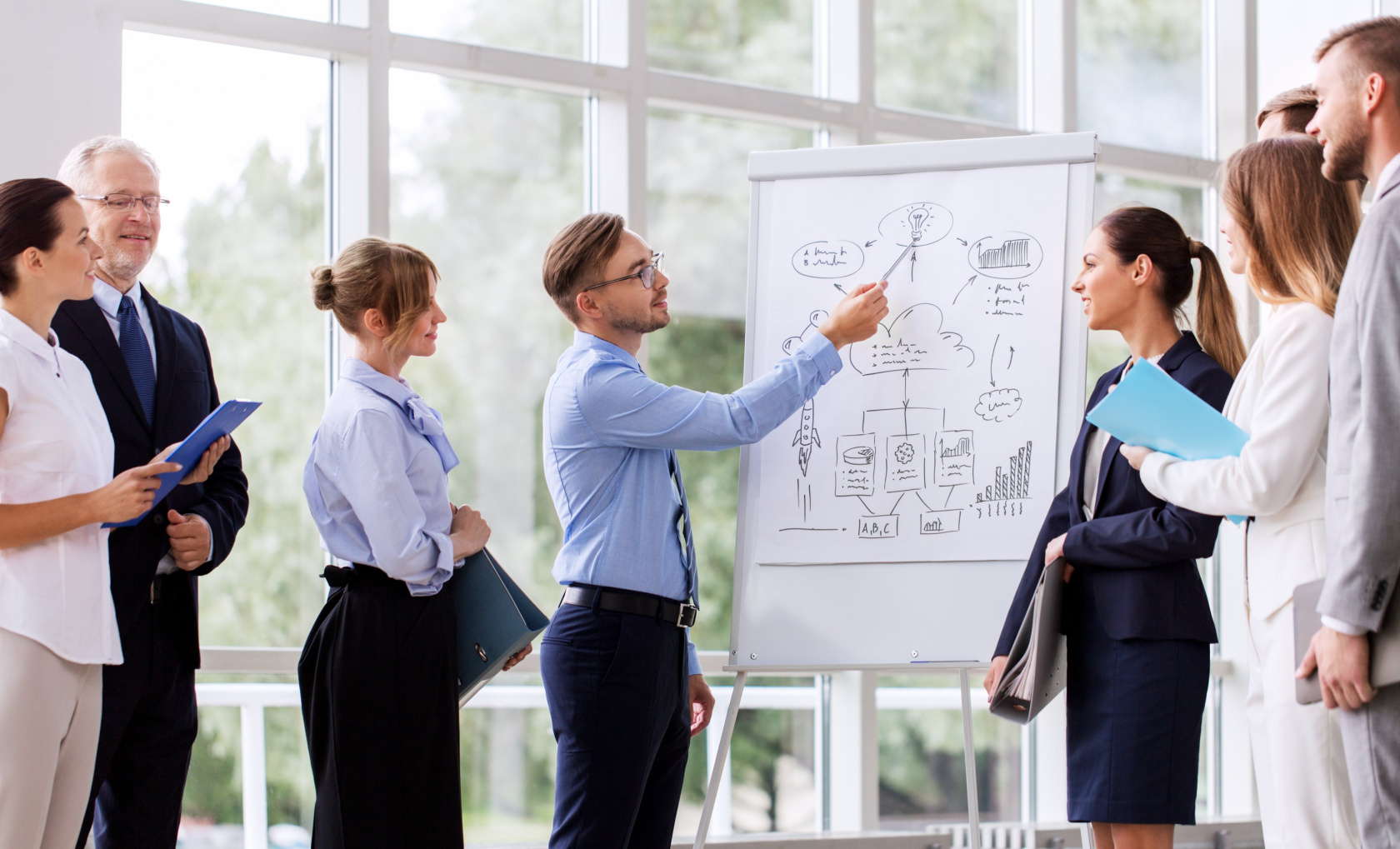 A man presenting a hand-drawn diagram on a flip chart to a group of colleagues in a bright atrium