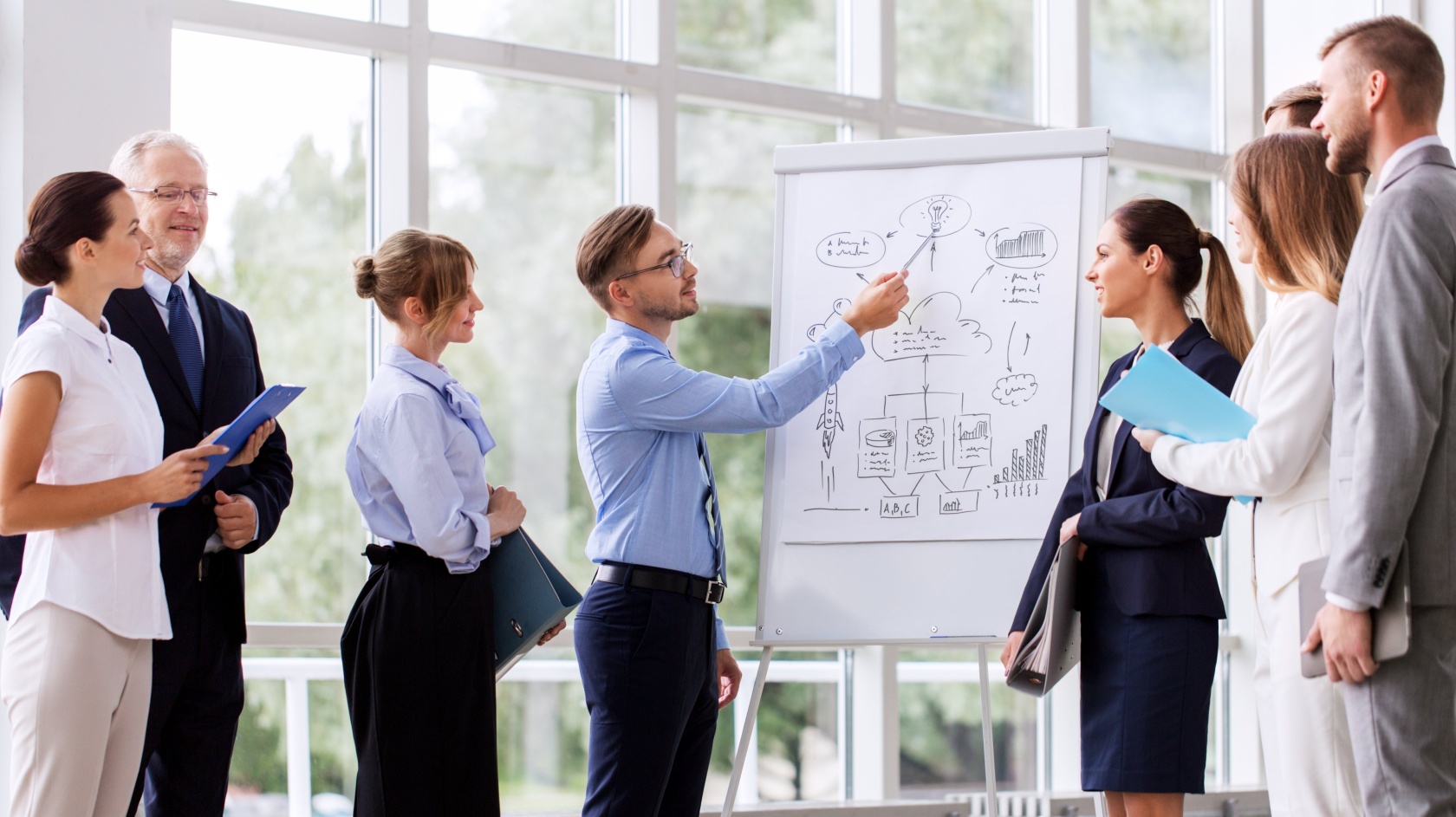 A man presenting a hand-drawn diagram on a flip chart to a group of colleagues in a bright atrium