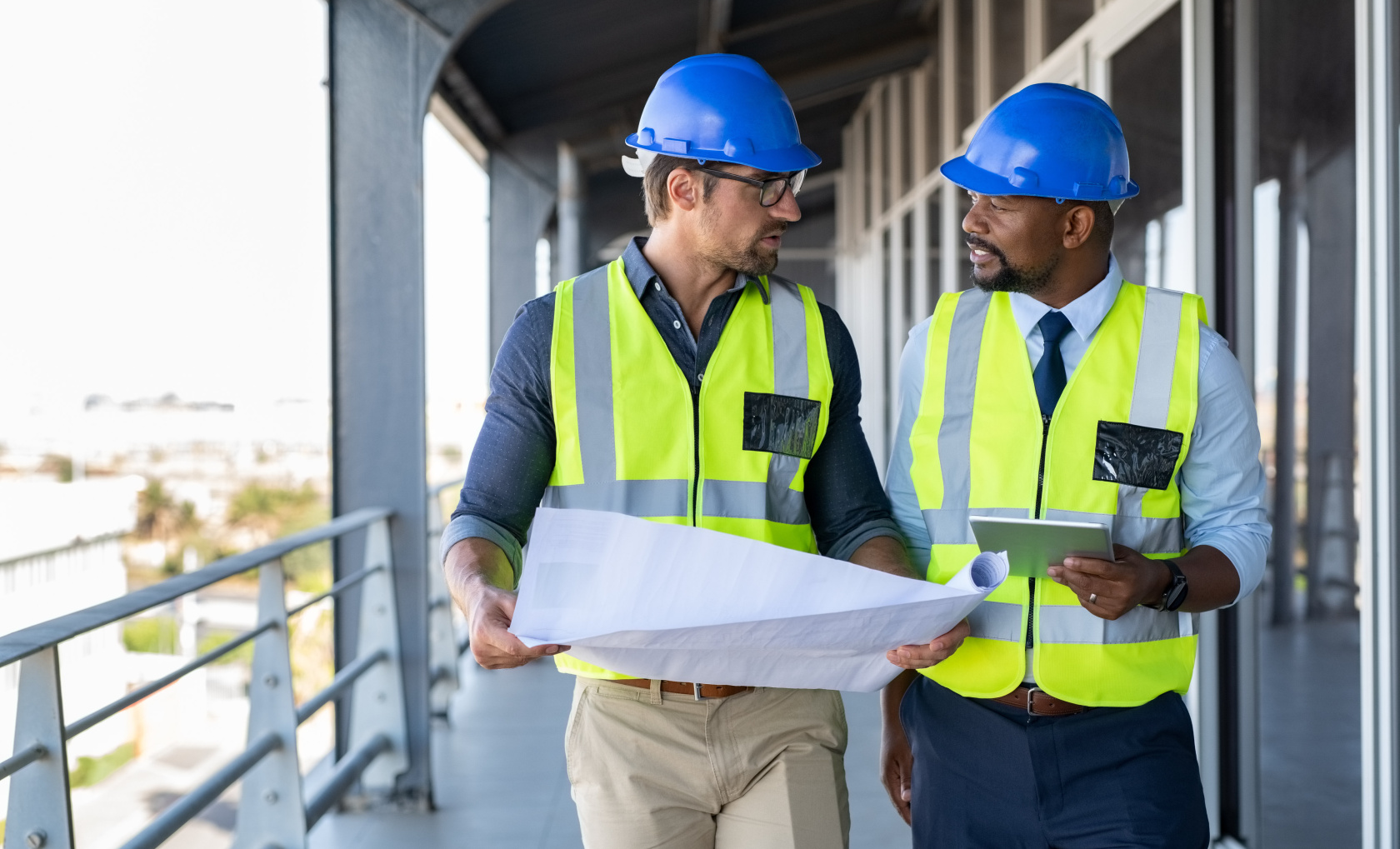 Two construction professionals in hard hats and high-visibility vests reviewing an unrolled building plan on an open construction floor, one holding a tablet