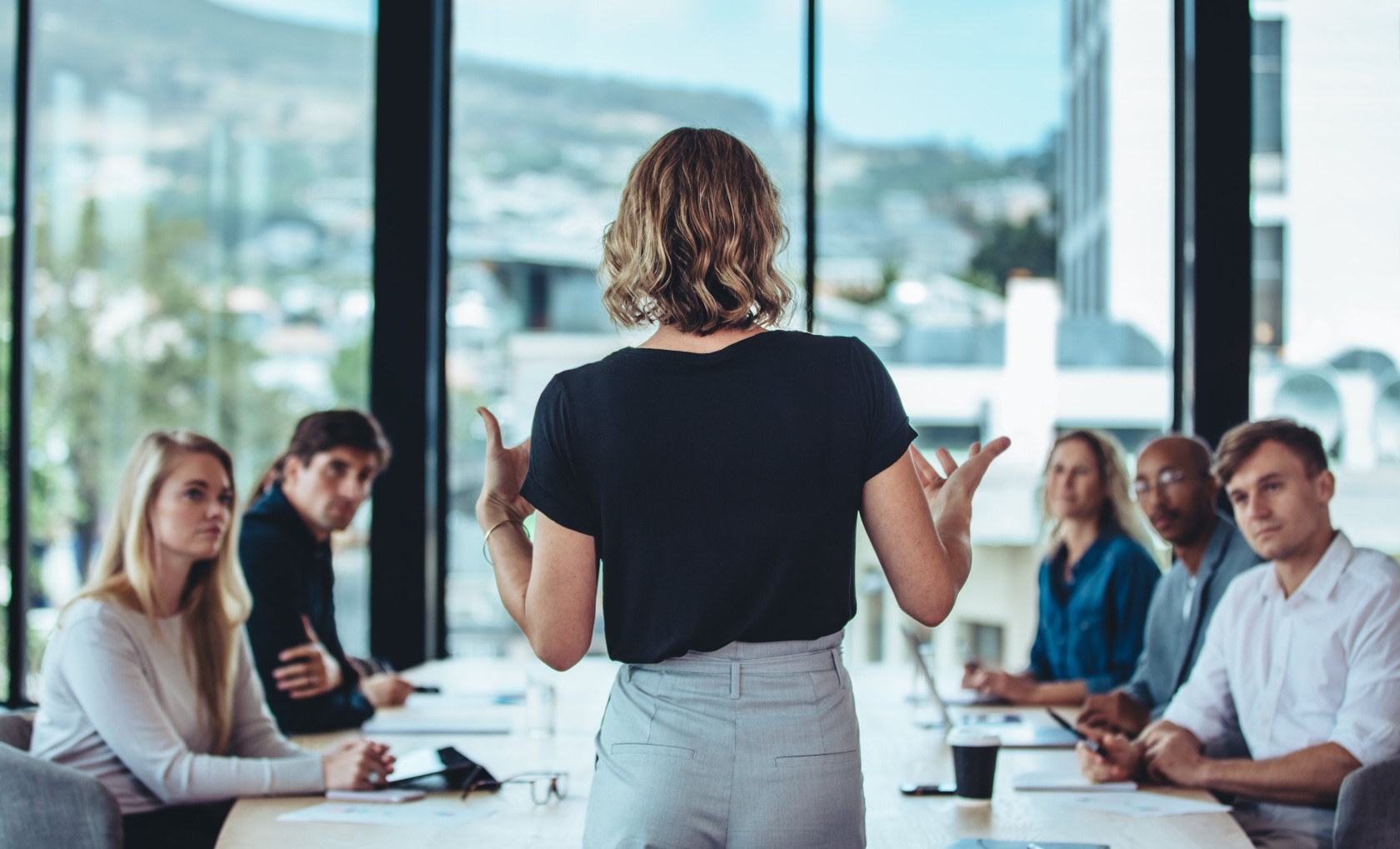 A woman standing with her back to the camera presenting to a seated team in a meeting room with mountain views