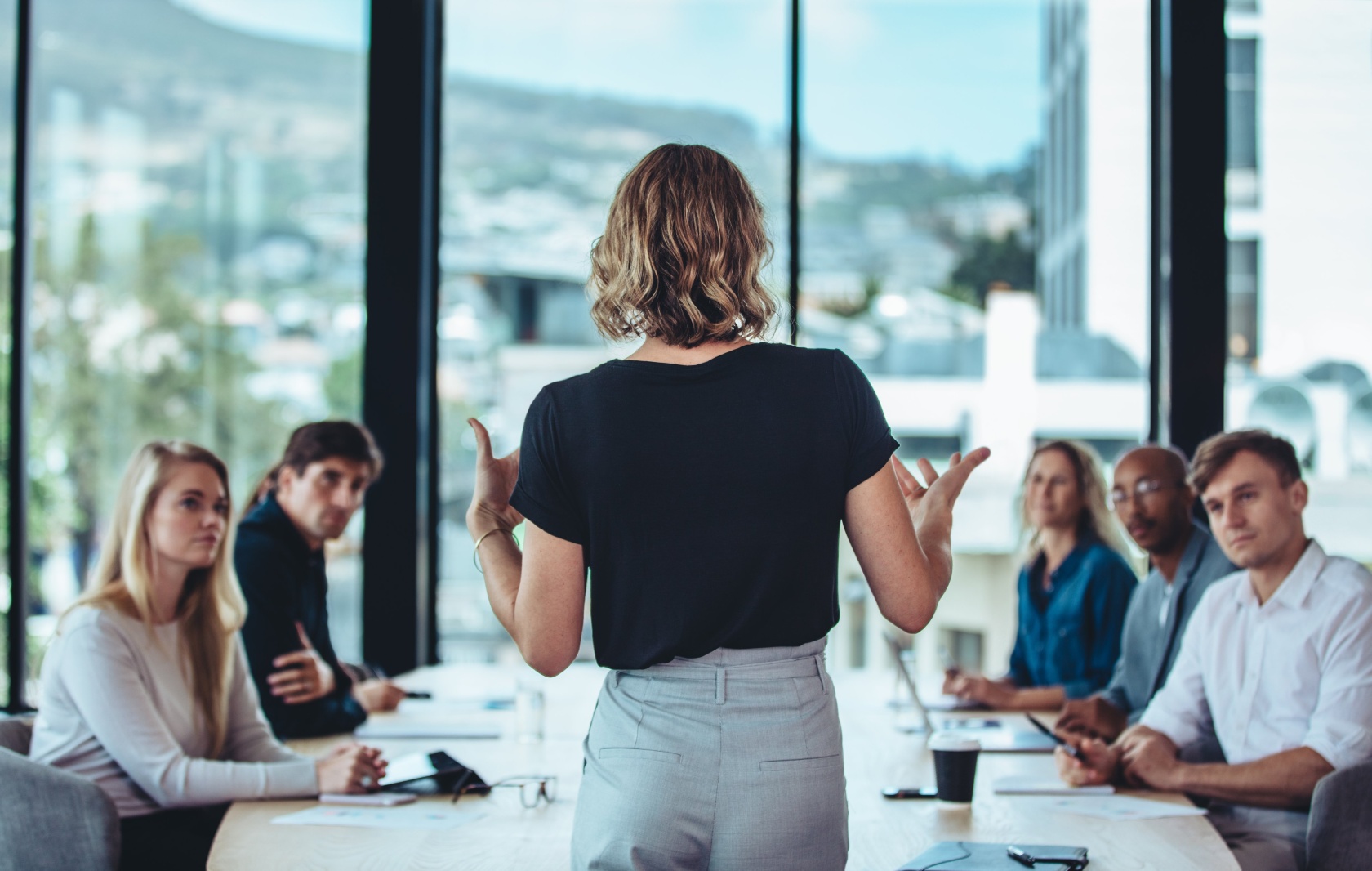A woman standing with her back to the camera presenting to a seated team in a meeting room with mountain views