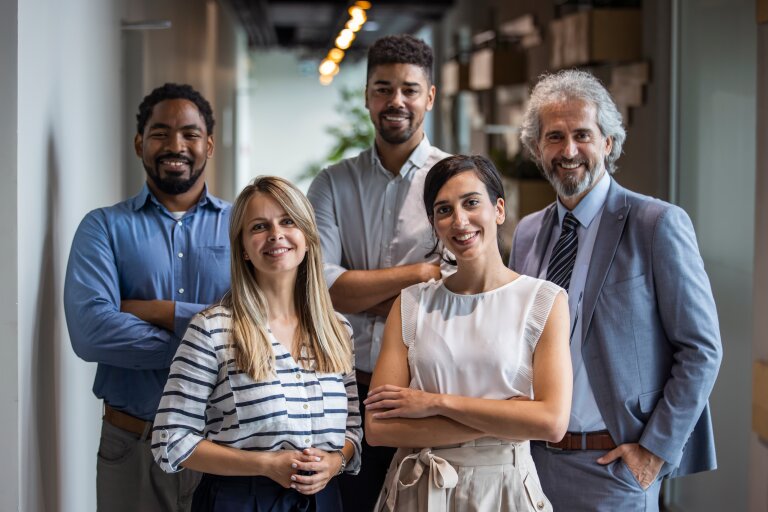 Five colleagues of different ages stand together in an office corridor and smile at the camera
