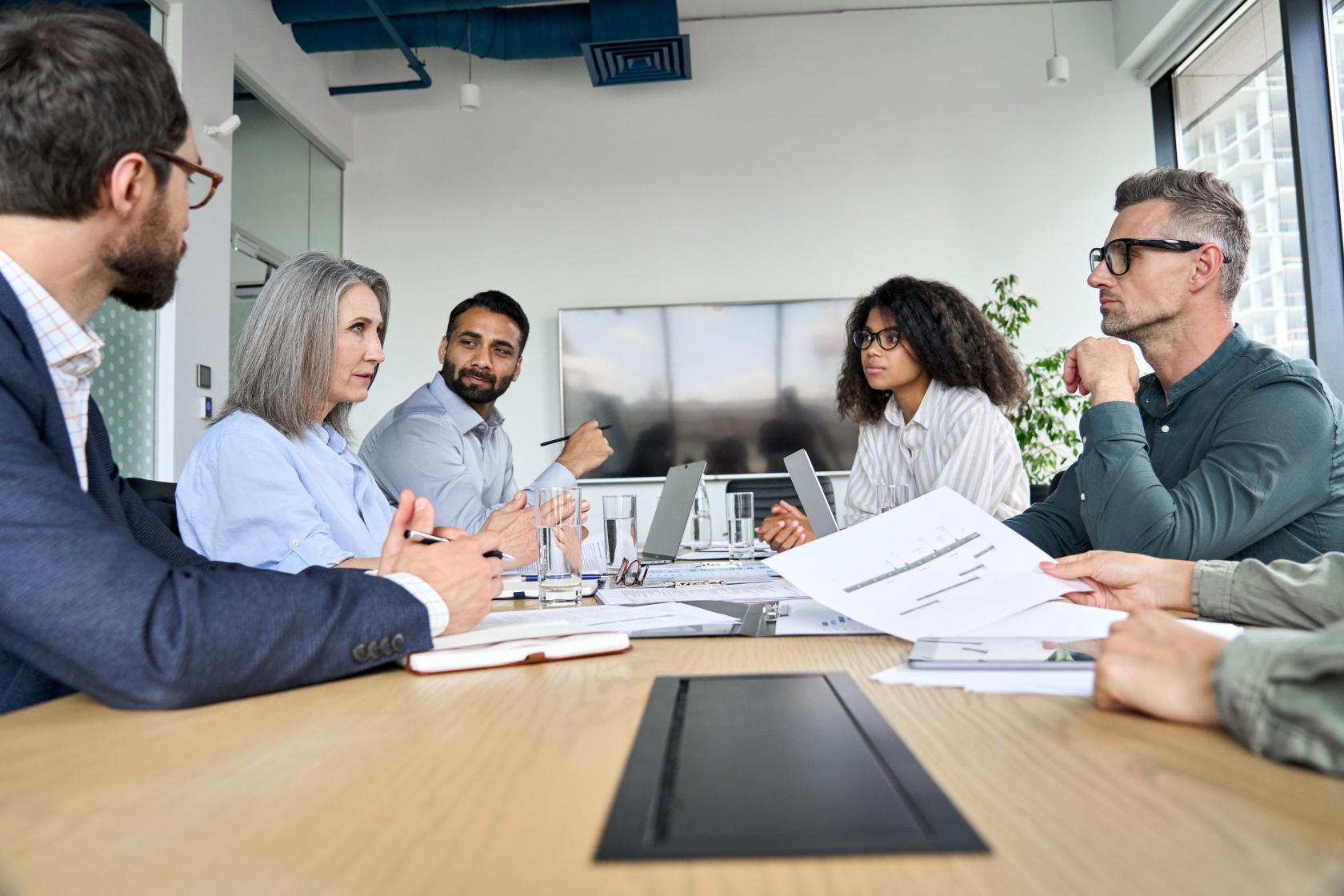 Six colleagues seated around a large meeting table in a bright office, discussing a printed document