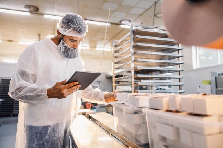 A worker in protective clothing and hairnet using a tablet beside packaged goods on a food production line