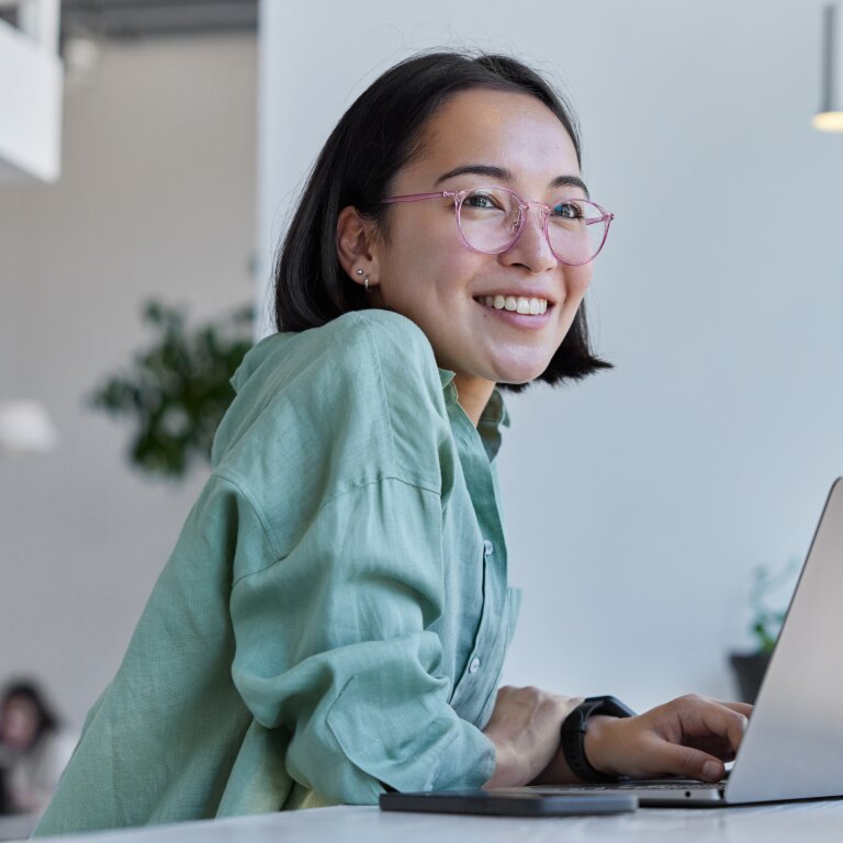 A smiling woman with glasses works at a laptop in a bright, open-plan office