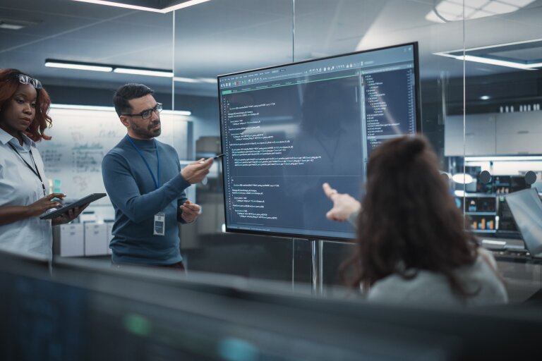 Three IT specialists in a control room discussing code shown on a large wall screen