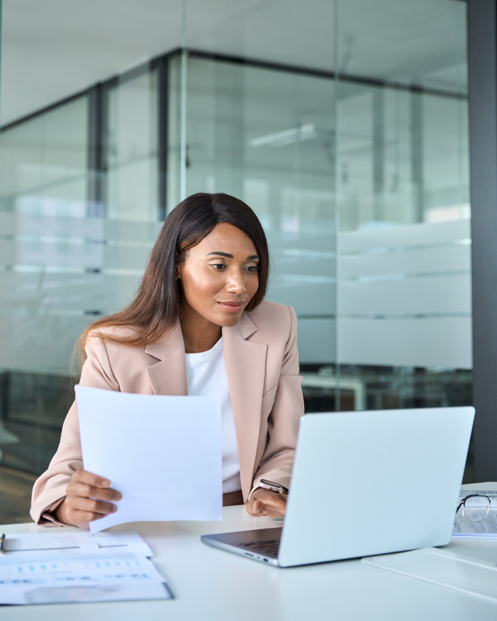 A woman in a pale blazer at a laptop in a glass-walled office, reading a printed report