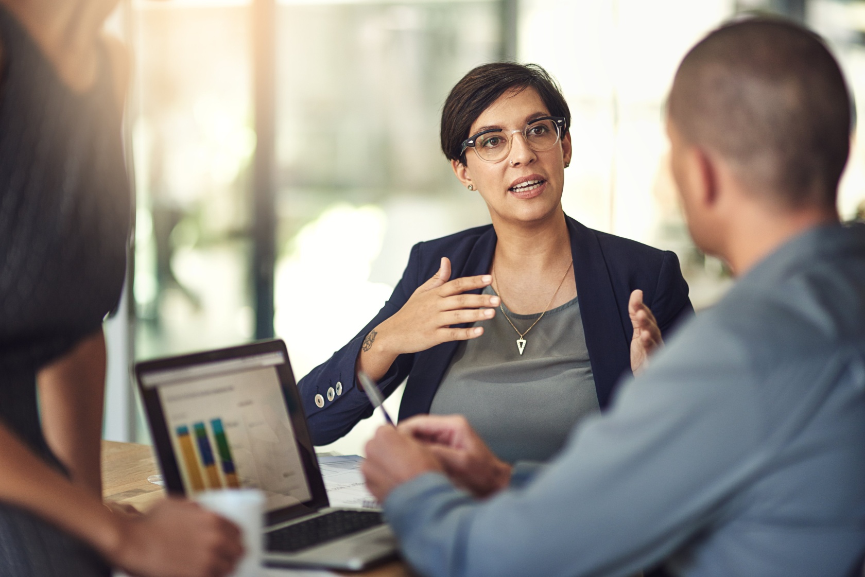 A woman in a blazer explaining something with her hands to a colleague across a laptop in a sunlit office
