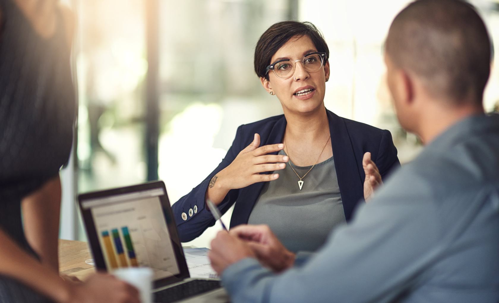 A woman in a blazer explaining something with her hands to a colleague across a laptop in a sunlit office