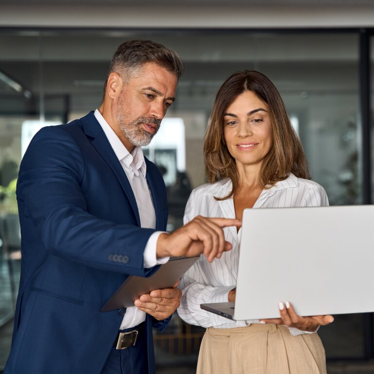 A man in a blue suit points at a laptop that a colleague is holding as the two review something together in an office