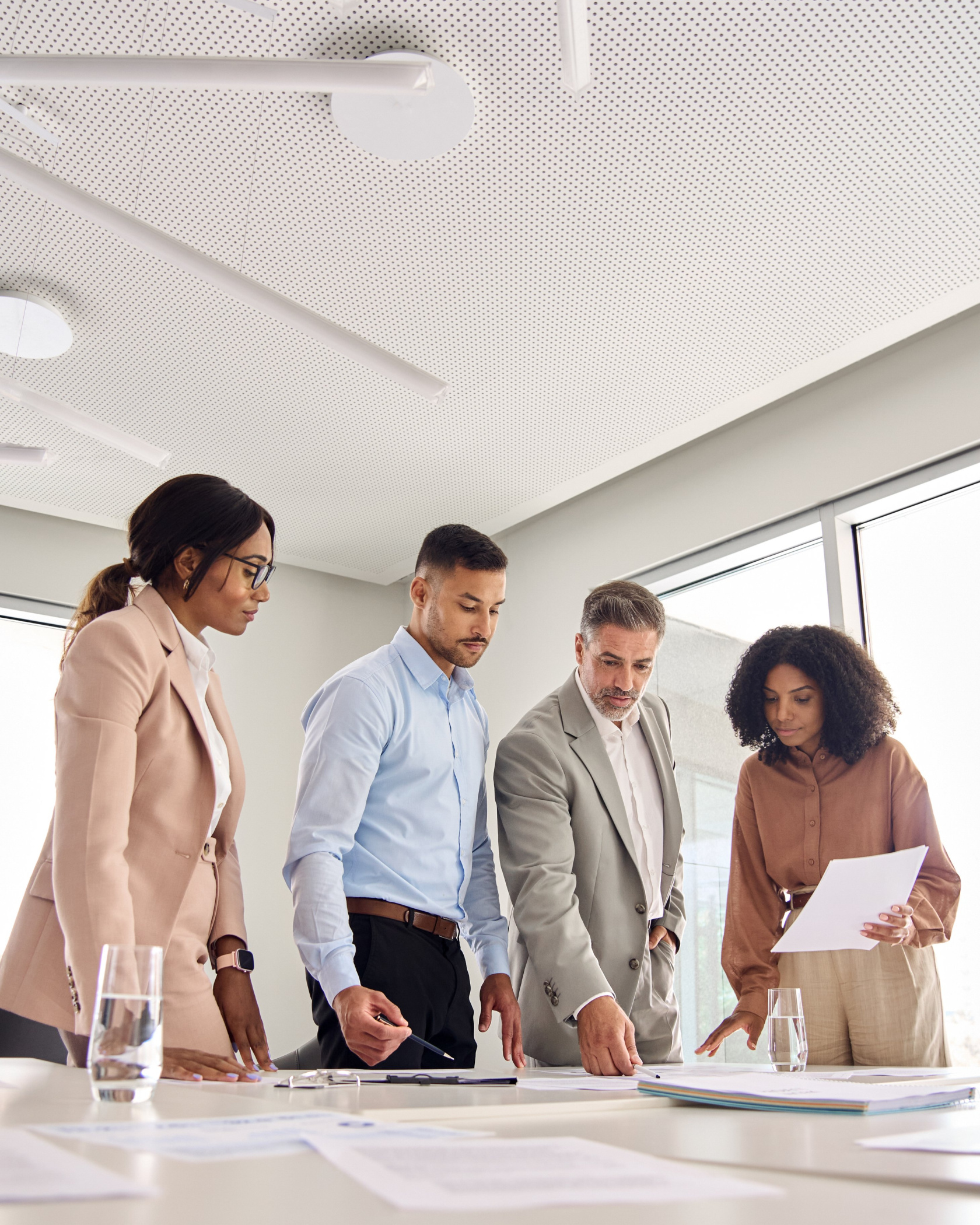 Four colleagues standing around a white conference table, leaning over printed documents in a bright meeting room