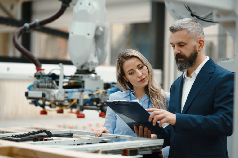 A man and a woman with a clipboard inspecting a robotic arm on a production line