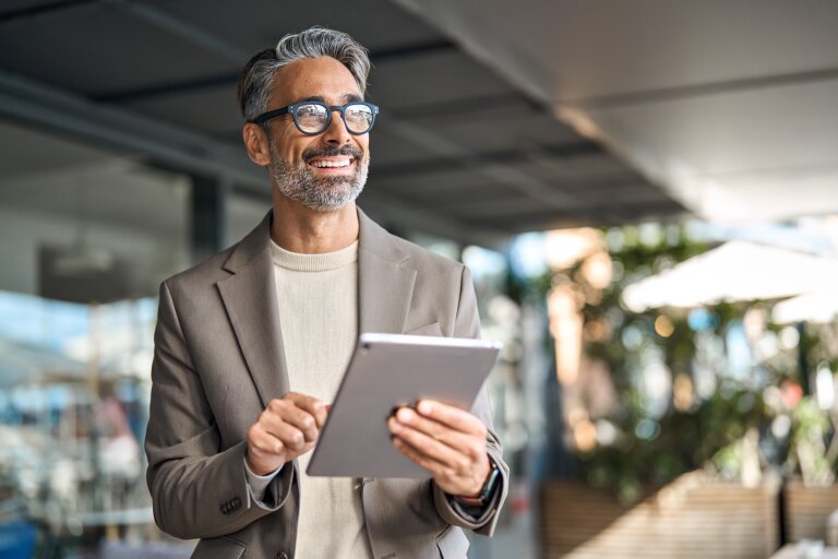 A smiling man with grey hair and glasses in a light blazer stands holding a tablet
