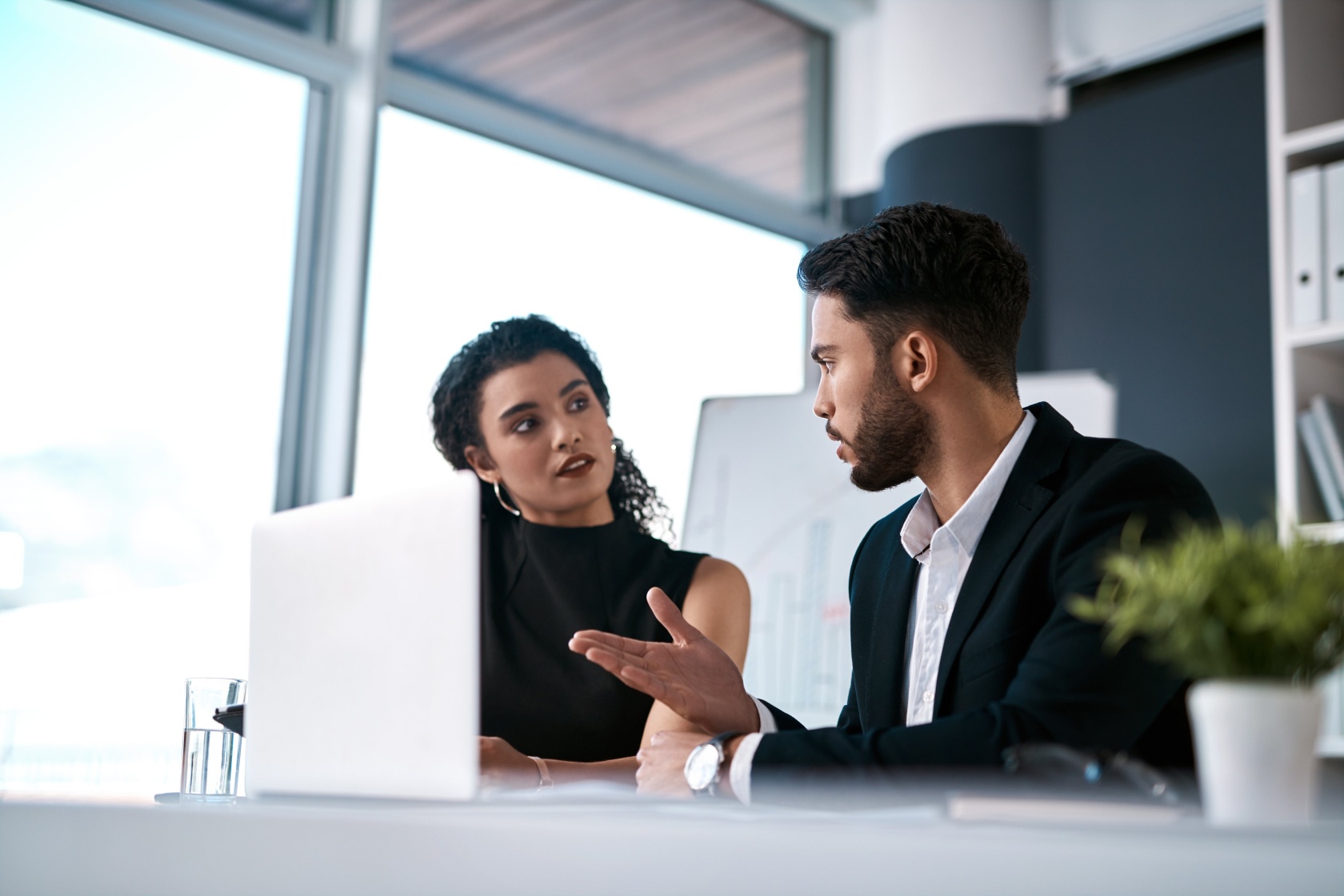 A woman and a man in conversation across a desk with a laptop between them in a bright office
