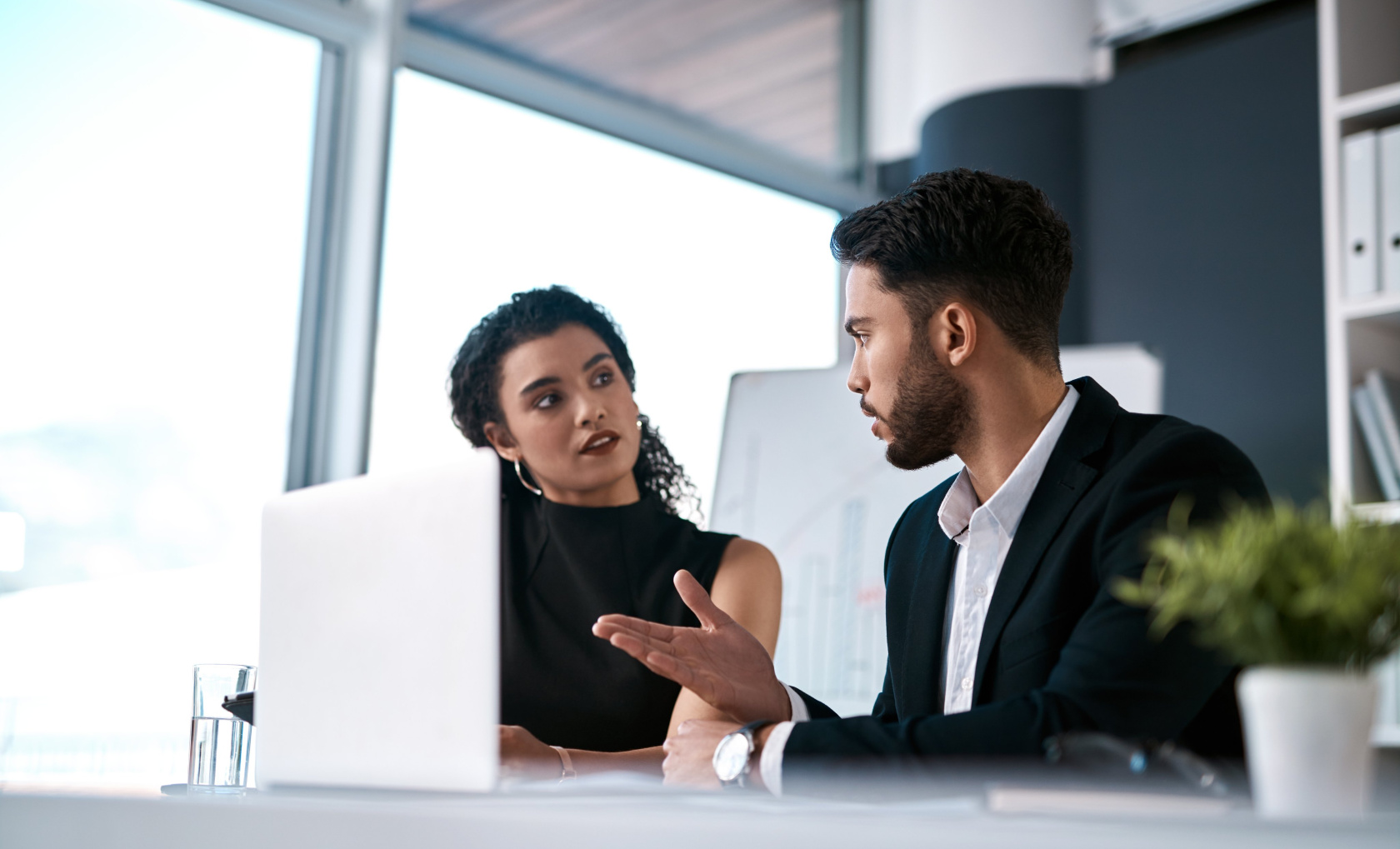 A woman and a man in conversation across a desk with a laptop between them in a bright office